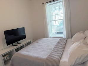 A bedroom with a bed, a television on a stand, a window with curtains, and a view of the outside. - Perfect 1-bedroom apartment (Doraville)