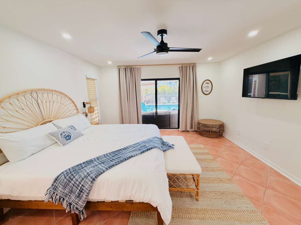 Ein Schlafzimmer mit einem Bett, einem Deckenventilator, einem Fenster mit Vorhängen und einem an der Wand montierten Fernseher.