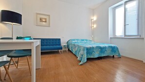 A small, well-lit room with a bed, a desk, and a chair. There is a window with blinds, a lamp, and a framed picture on the wall. - The Two Olive Trees - North (Campo nell'Elba)