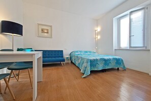 Ein kleiner, gut beleuchteter Raum mit einem Bett, einem Schreibtisch und einem Stuhl. Es gibt ein Fenster mit Jalousien, eine Lampe und ein gerahmtes Bild an der Wand.