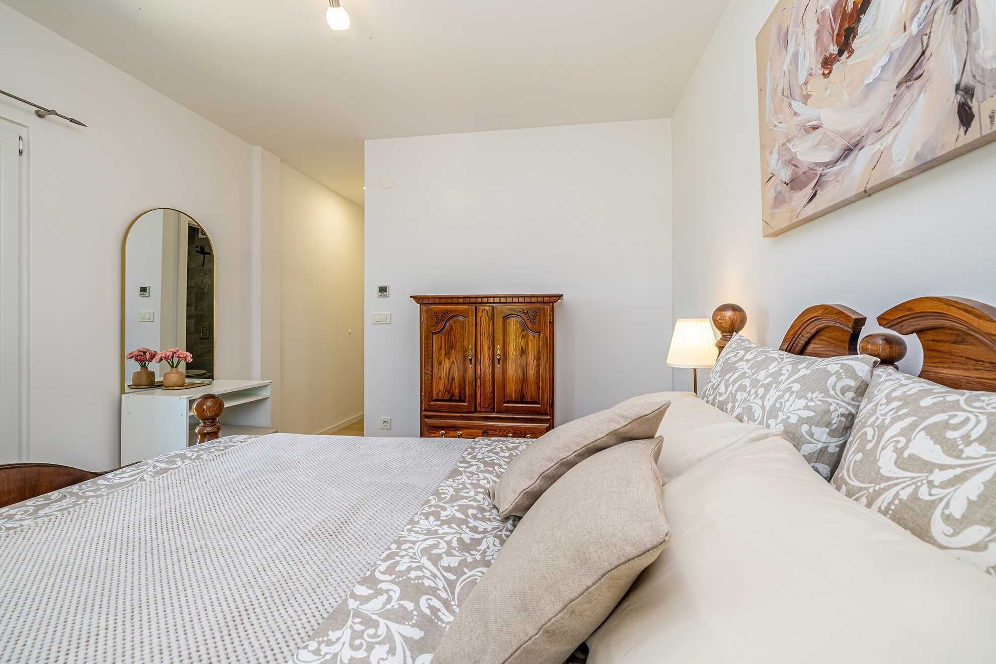A bedroom with a bed, a wooden wardrobe, a mirror, and a painting on the wall.