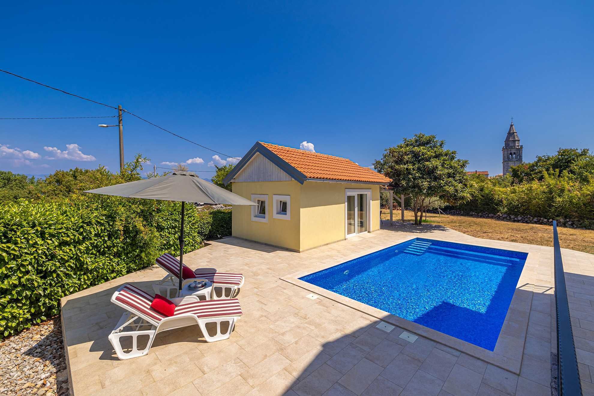 A pool area with lounge chairs and a small building, likely a house or guest house, with a clear view of a tower in the background.