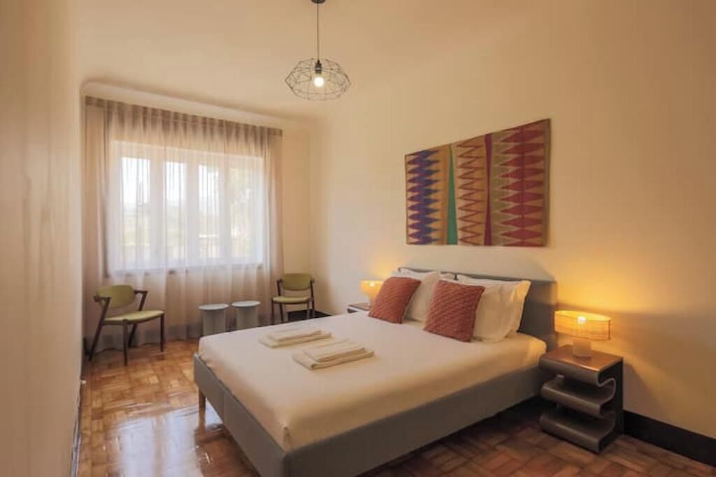 Habitación de hotel con cama, escritorio, silla, ventana con cortinas, una obra de arte en la pared y una lámpara colgante.