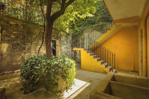 A courtyard with a staircase leading to an upper level, surrounded by stone walls and greenery.