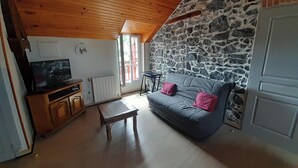 Ein gemütliches Wohnzimmer mit Steinwand, Holzboden, einer grauen Couch mit rosafarbenen Kissen, einem Holztisch und einem Fernseher auf einem Holzschrank.
