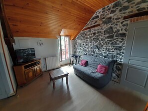 A cozy living room with a stone wall, a wooden ceiling, a grey sofa with pink cushions, a wooden coffee table, and a television on a wooden cabinet. - One-Bedroom Apartment for Rent in the Heart of the Basque Country (Cambo-les-Bains)
