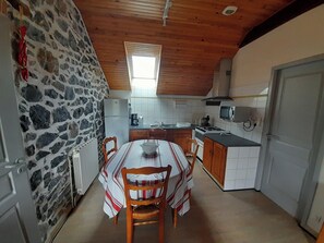 A kitchen with a stone wall, wooden ceiling, a dining table with chairs, and a window. - One-Bedroom Apartment for Rent in the Heart of the Basque Country (Cambo-les-Bains)