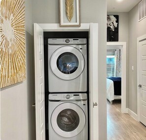 A laundry room with two Samsung washing machines, a decorative leaf, and a framed artwork on the wall. - 2 Beds Near Downtown, Stockyards, Zoos, Sundance, 6 Flags,AT&T Stadium, Botanic (Fort Worth)