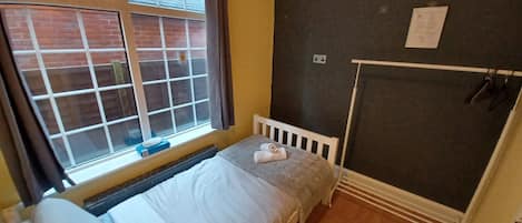 Une petite pièce avec un lit simple, une fenêtre avec des rideaux, un radiateur et un chauffage fixé au mur.