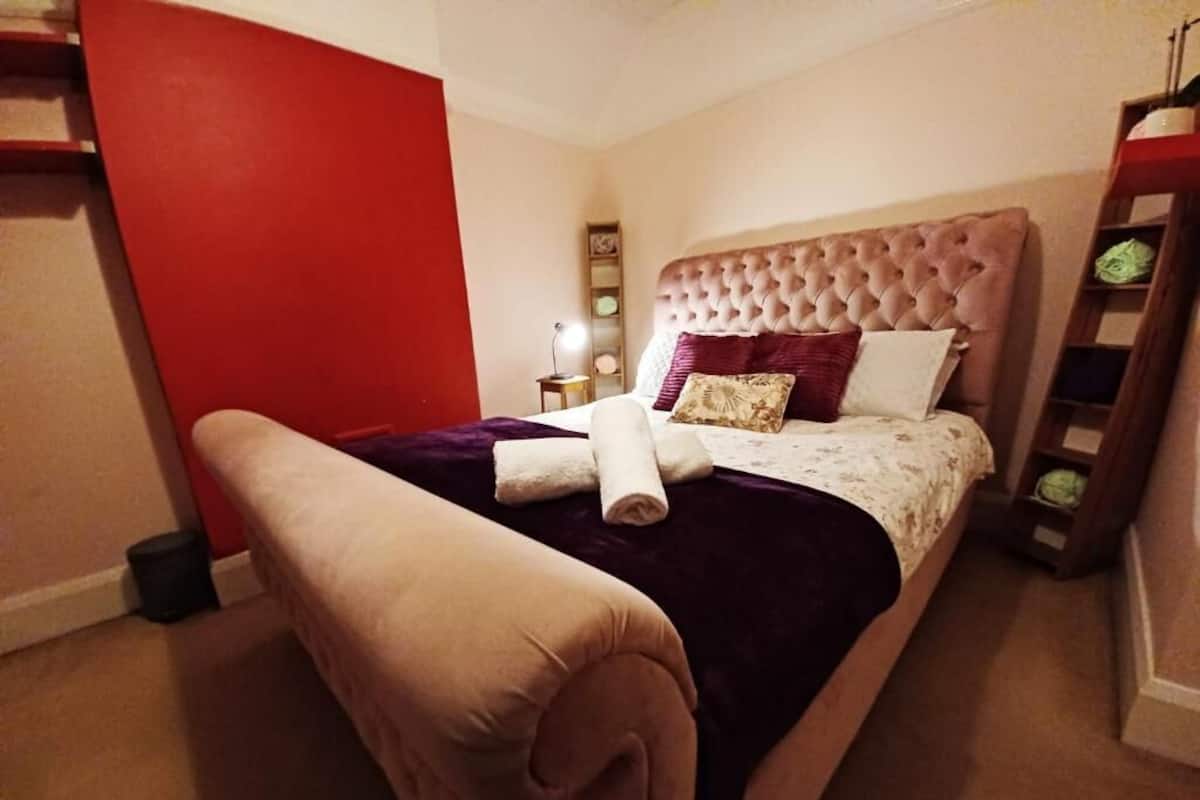 Een net opgemaakt bed met een gestuufd hoofdbord, een nachtkastje met een lamp, en een rode accentmuur.