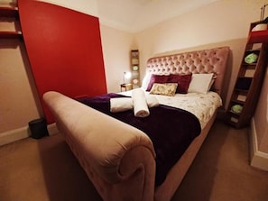 Een net opgemaakt bed met een gestuufd hoofdbord, een nachtkastje met een lamp, en een rode accentmuur.