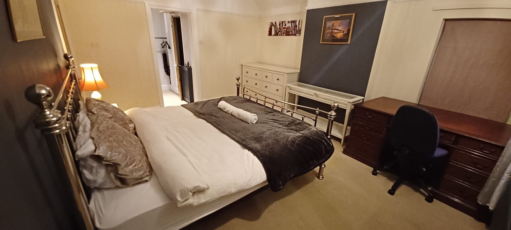 A bedroom with a bed, bedside table, dresser, desk, chair, and a door leading to another room.