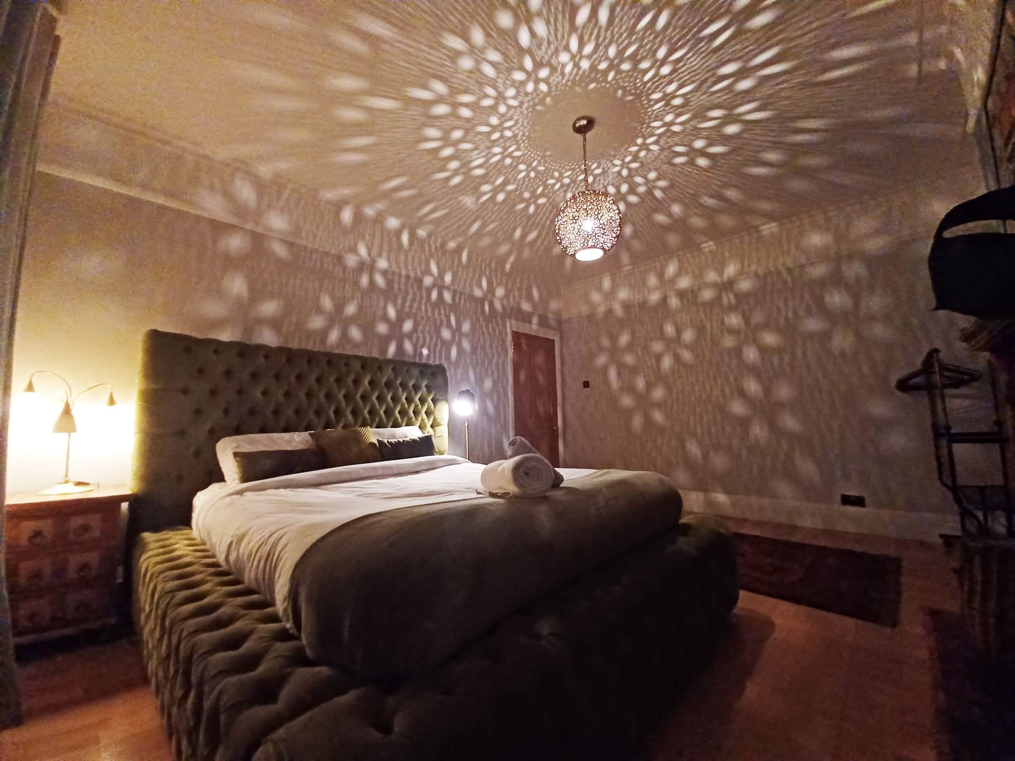 Une chambre à coucher avec une tête de lit capitonnée, un lit sur lequel est posée une serviette roulée, et un luminaire décoratif au plafond.