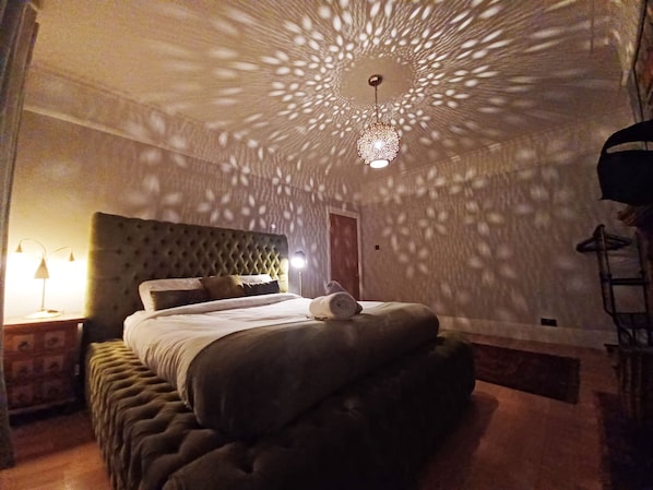 A bedroom with a tufted headboard, a bed with a rolled towel, and a decorative ceiling light. - The Gemstone Manor (Watford)