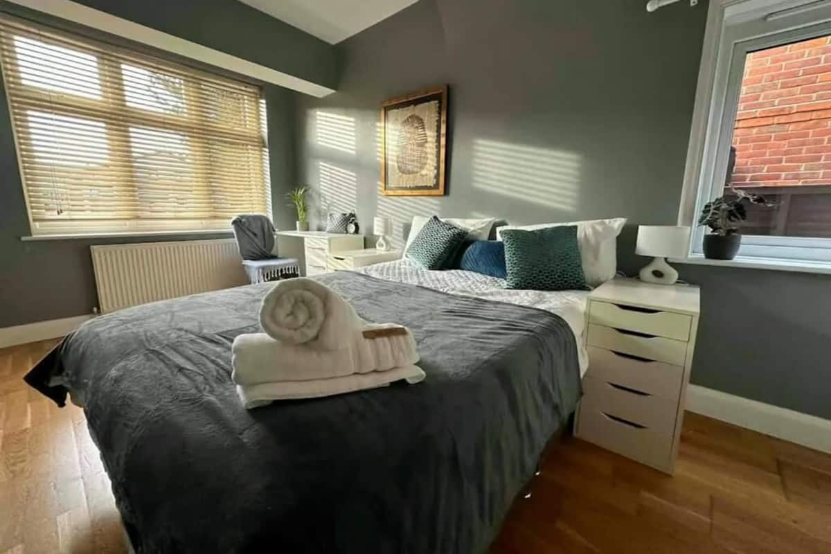 Een slaapkamer met een bed, bureau, stoel, radiator, raam met jaloezieën en een plant in pot.