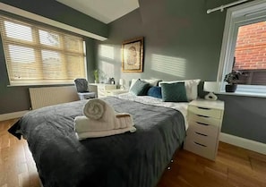 Een slaapkamer met een bed, bureau, stoel, radiator, raam met jaloezieën en een plant in pot.