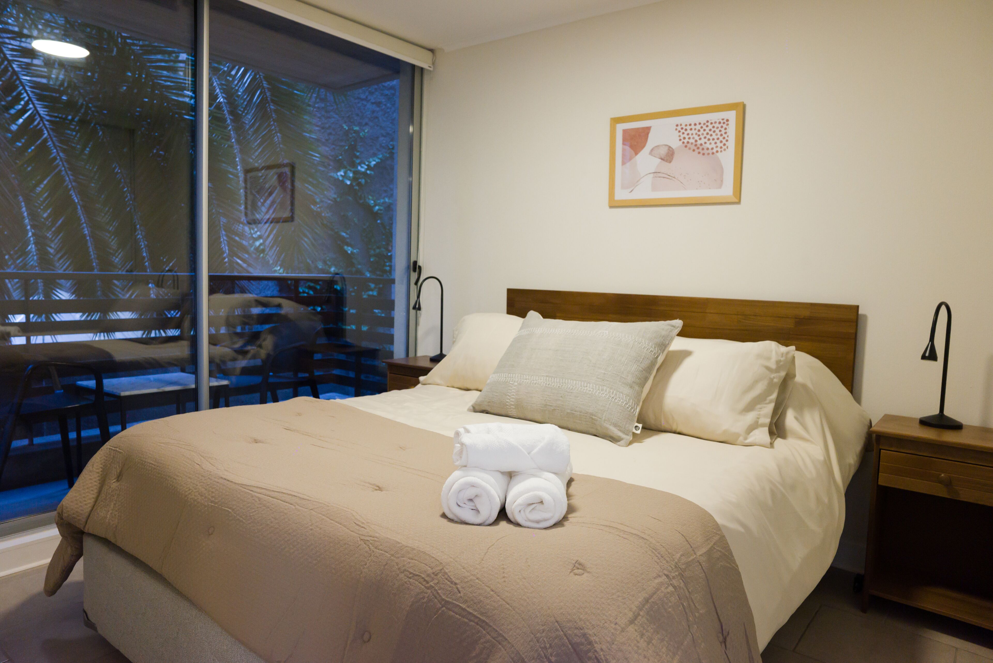 Una habitación de hotel con una cama, una mesita de noche, una lámpara, una ventana con vista a palmeras y un cuadro en la pared.