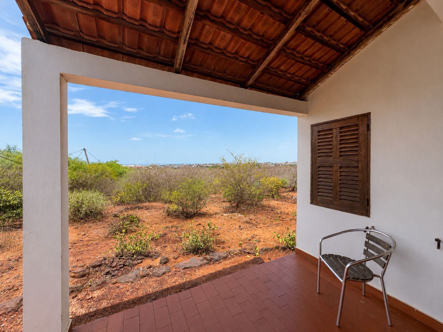 Una habitación con vista a un paisaje desértico, una ventana con contraventanas y una silla de metal.