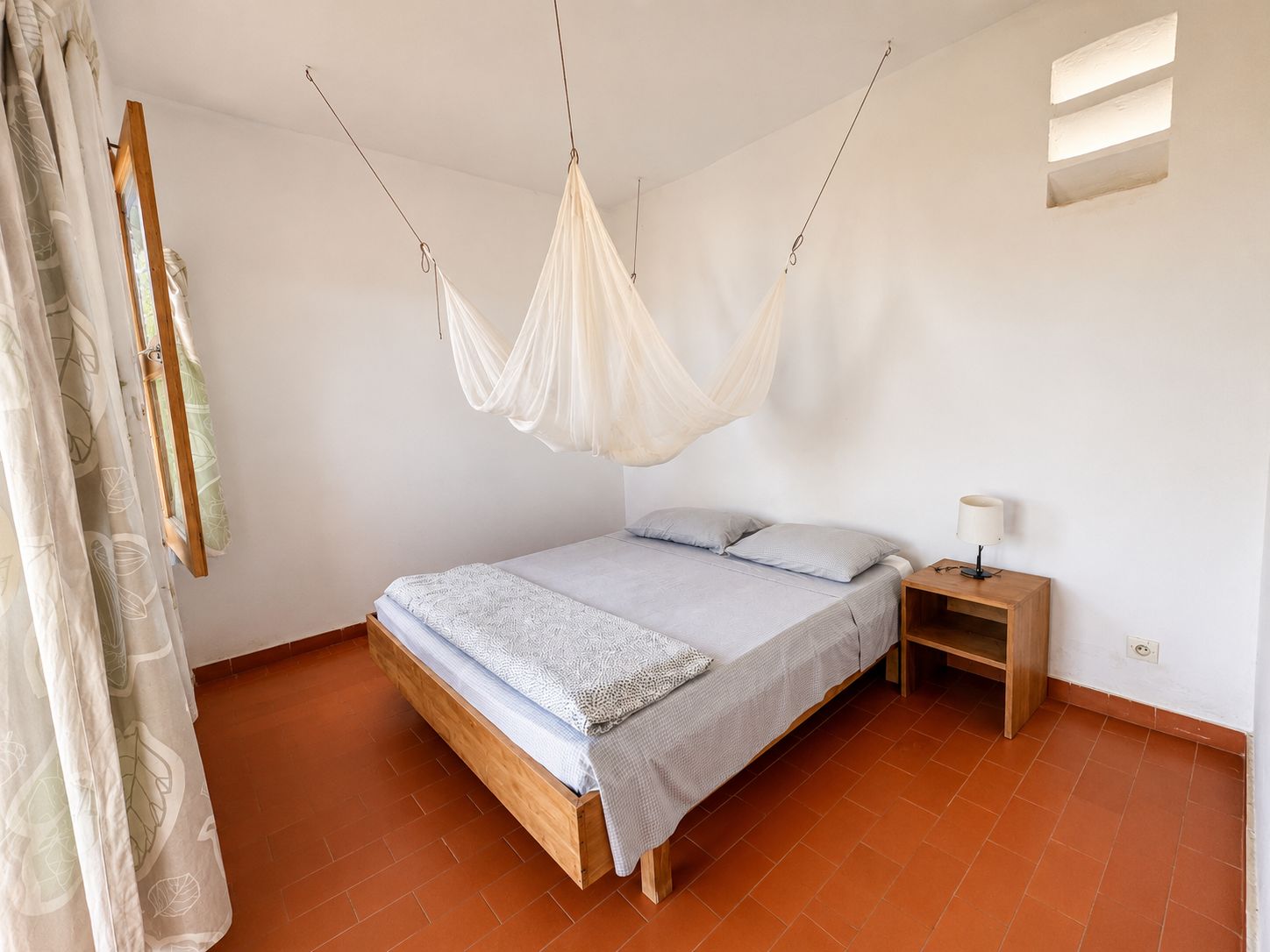 Un dormitorio con cama, mesita de noche, dosel colgante y ventana con persianas.