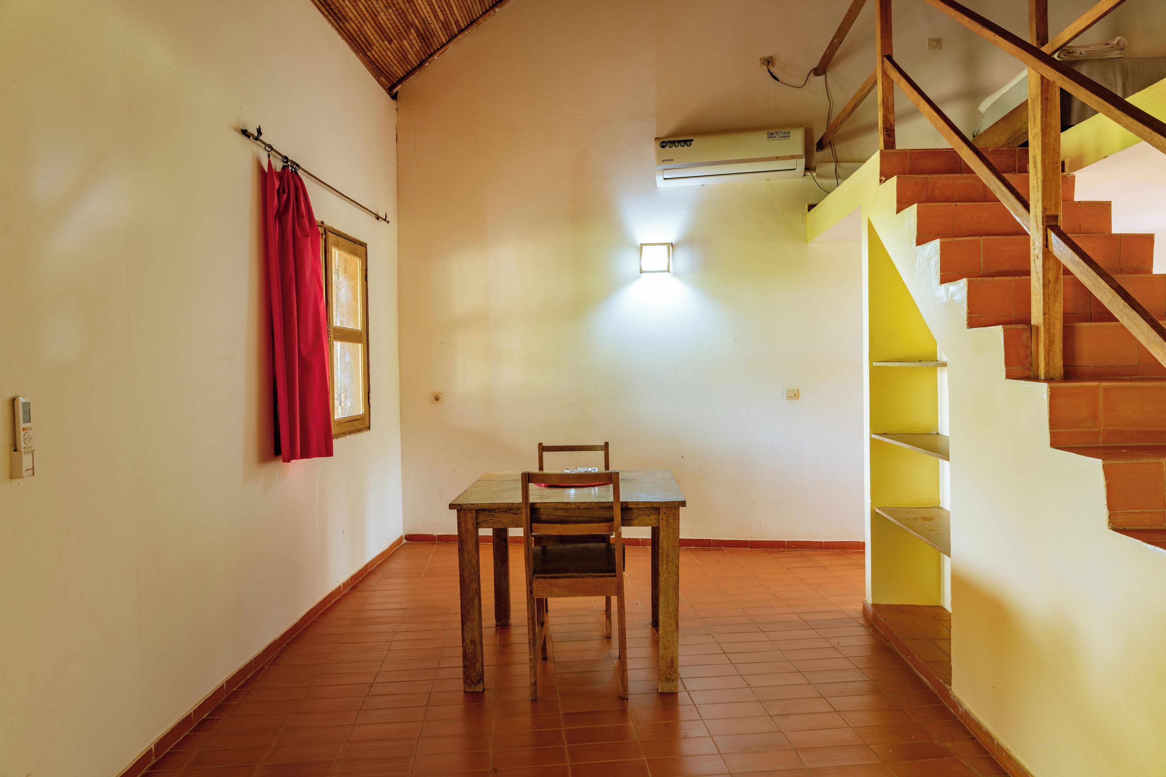 Una habitación con una mesa y una silla de madera, una cortina roja, una ventana y una escalera con escalones amarillos.