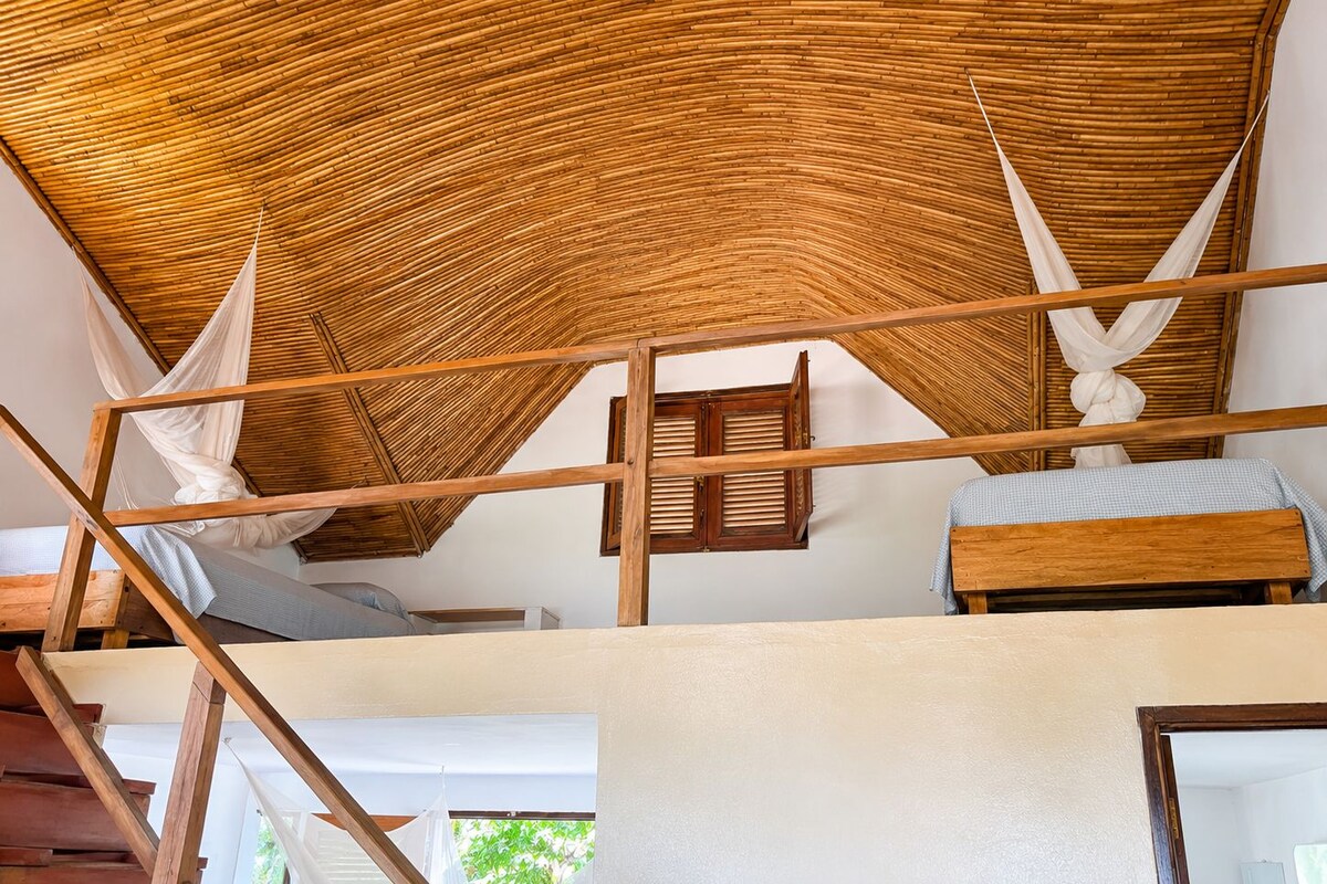 A room with a high ceiling, wooden beams, a bed with a white bedspread, and a window with wooden shutters.