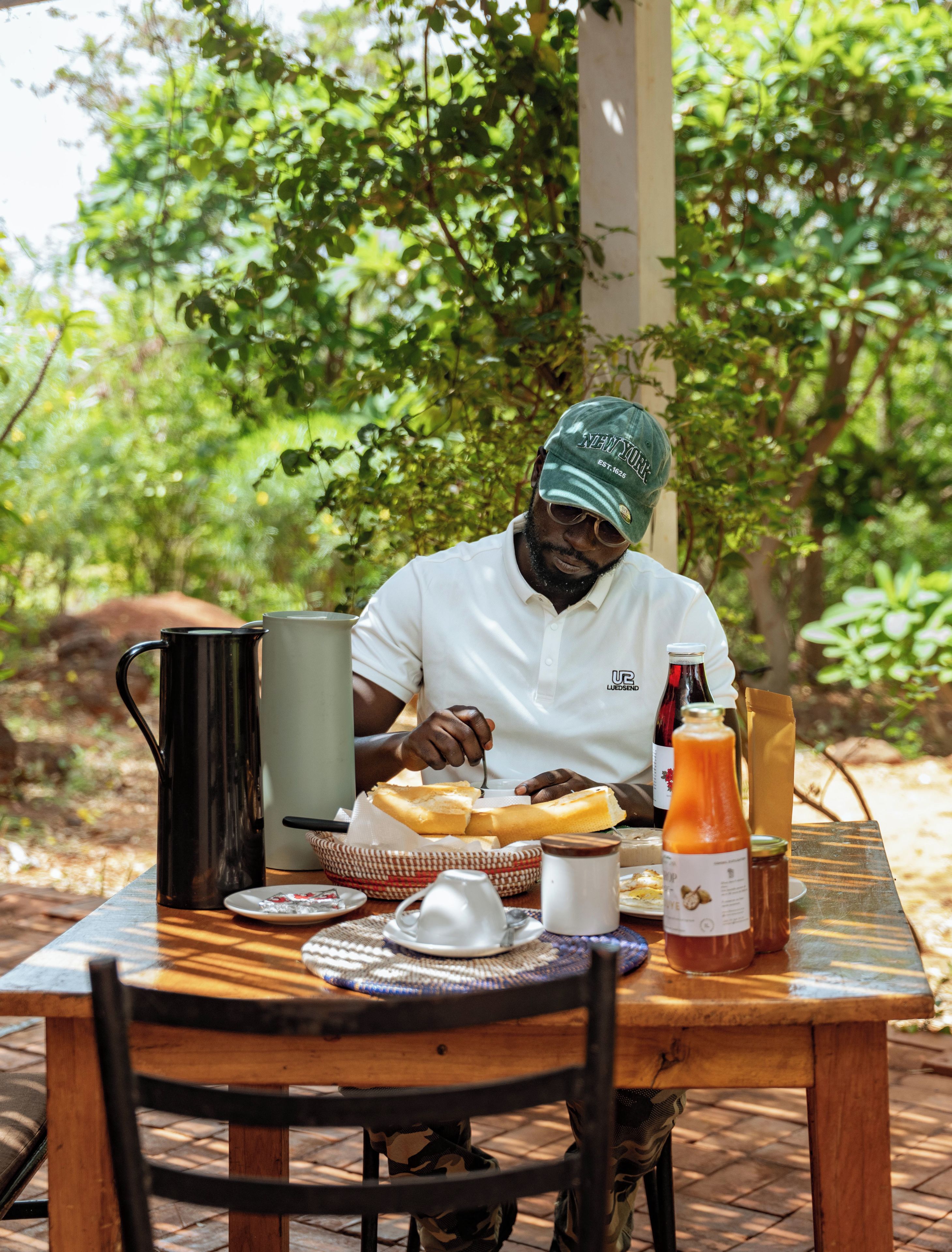 A person seated at an outdoor table with a meal, a black coffee pot, a bottle of juice, and a bottle of wine.