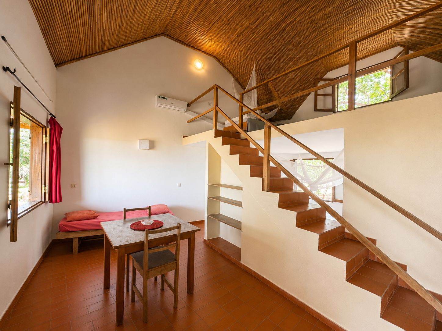 Amplia habitación con escalera de madera, comedor con mesa y sillas, y ventana con vista a follaje.