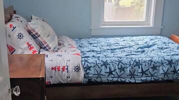 Una cama individual con colcha azul y blanca estampada, cabecera de madera y ventana con vista a árboles.