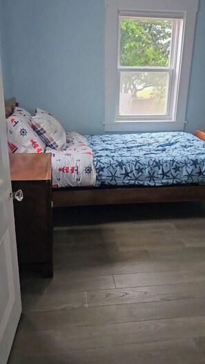 A single bed with a blue and white patterned bedspread, a wooden headboard, and a window with a view of trees.