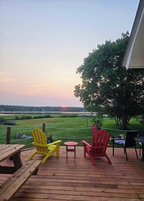 A deck with wooden benches and Adirondack chairs, overlooking a grassy field and a sunset.