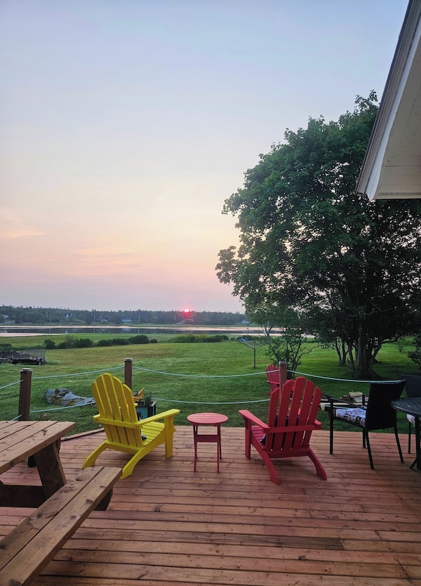 A deck with wooden benches and Adirondack chairs, overlooking a grassy field and a sunset.