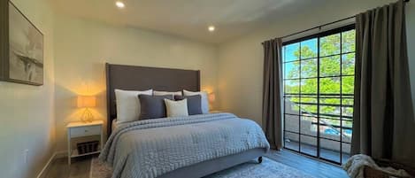 A bedroom with a large bed, a nightstand, a lamp, and a view of greenery through a window.