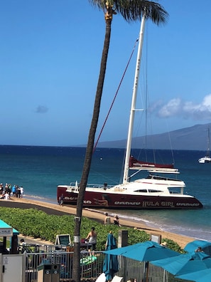 A beachside view with a sailboat named 'Hula Girl' anchored near the shore, a palm tree, and a clear blue sky.
