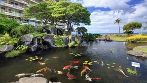 Ein Teich mit Koi-Karpfen und einem Wasserfall, umgeben von üppiger Vegetation, mit einem Hotel im Hintergrund.