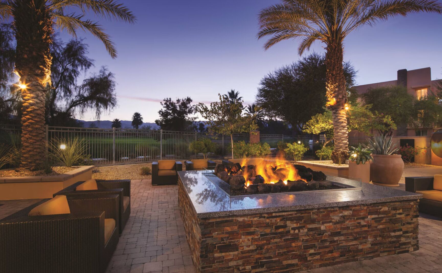 An outdoor fire pit area with seating, surrounded by palm trees and a brick wall.
