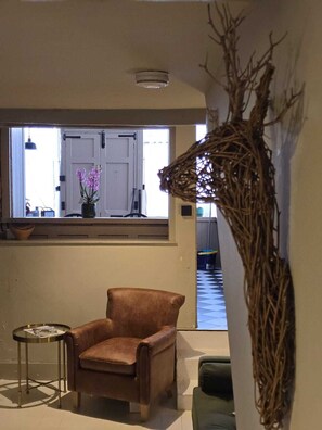 A room with a brown leather armchair, a small round table with a magazine, a large window with a view of the outside, and a decorative branch hanging on the wall.