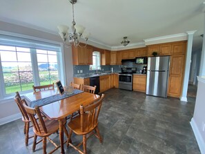 A modern kitchen with wooden cabinets, a stainless steel refrigerator, and a dining area with a wooden table and chairs. - PEI Cozy Clean Cottage with Hotub (Summerside)