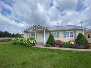 A two-story house with a front porch, a chimney, and a well-maintained lawn with various shrubs and flowers. - PEI Cozy Clean Cottage with Hotub (Summerside)