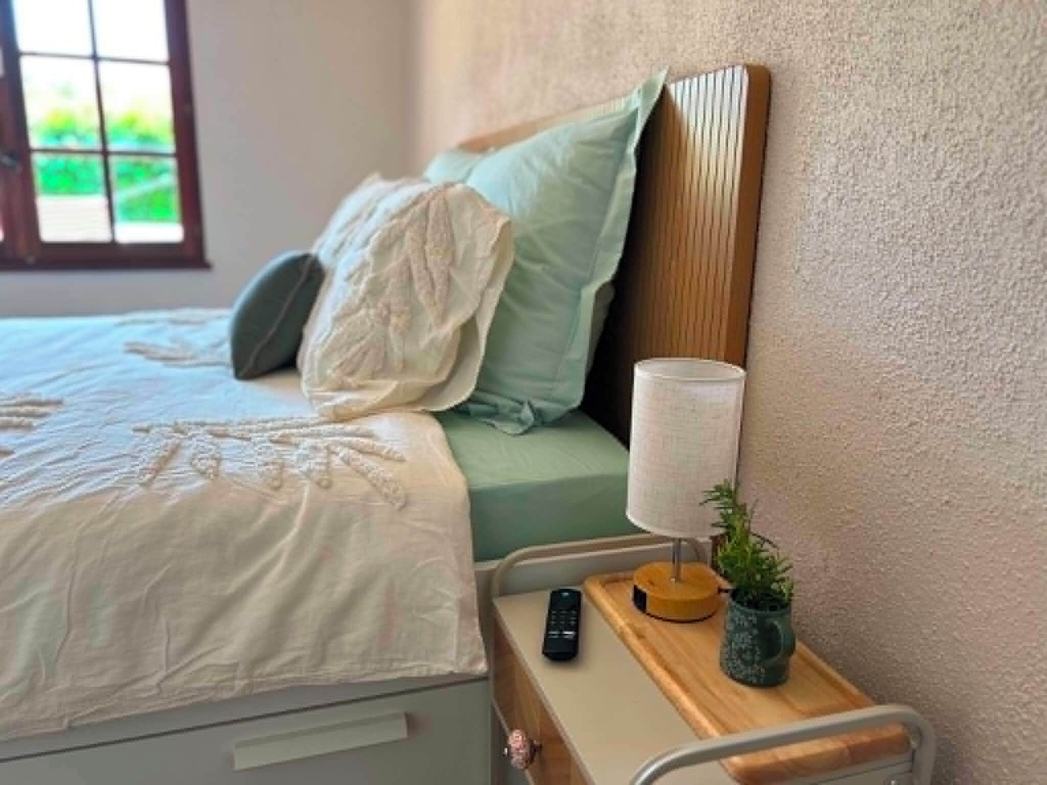Een bed met een witte dekbedovertrek en kussens, een bedlampje, een afstandsbediening en een kleine plant op een nachtkastje.