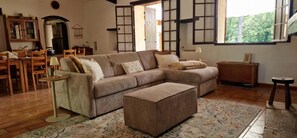 Uma sala de estar com sofá seccional, pufe, piso de madeira e vista pela janela.