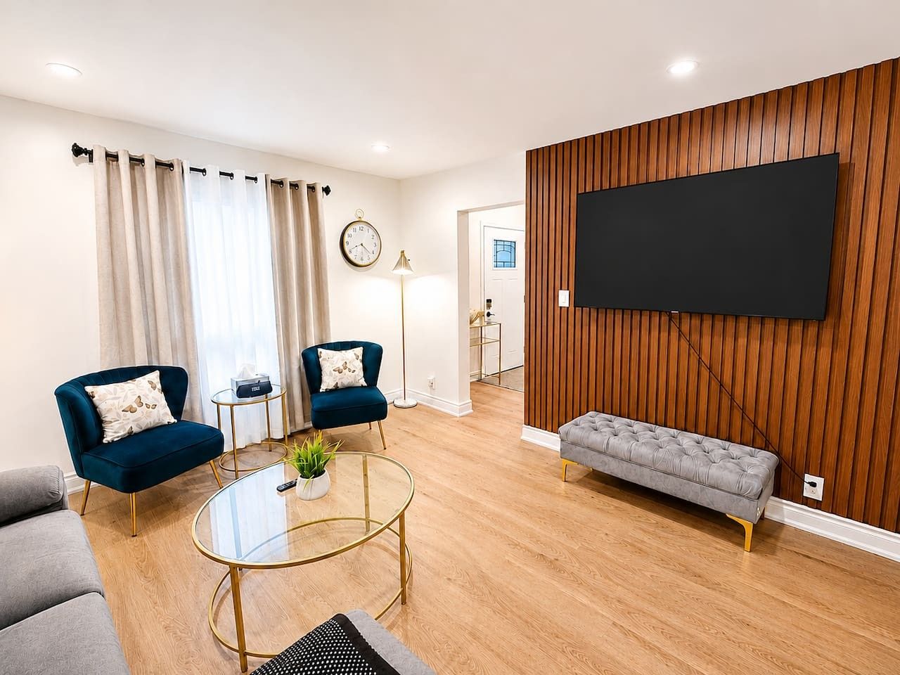 Un salon moderne avec un téléviseur à écran plat fixé à un mur en bois, deux fauteuils, un canapé, une table basse en verre et une table d’appoint avec une lampe.