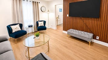 Un salon moderne avec un téléviseur à écran plat fixé à un mur en bois, deux fauteuils, un canapé, une table basse en verre et une table d’appoint avec une lampe.