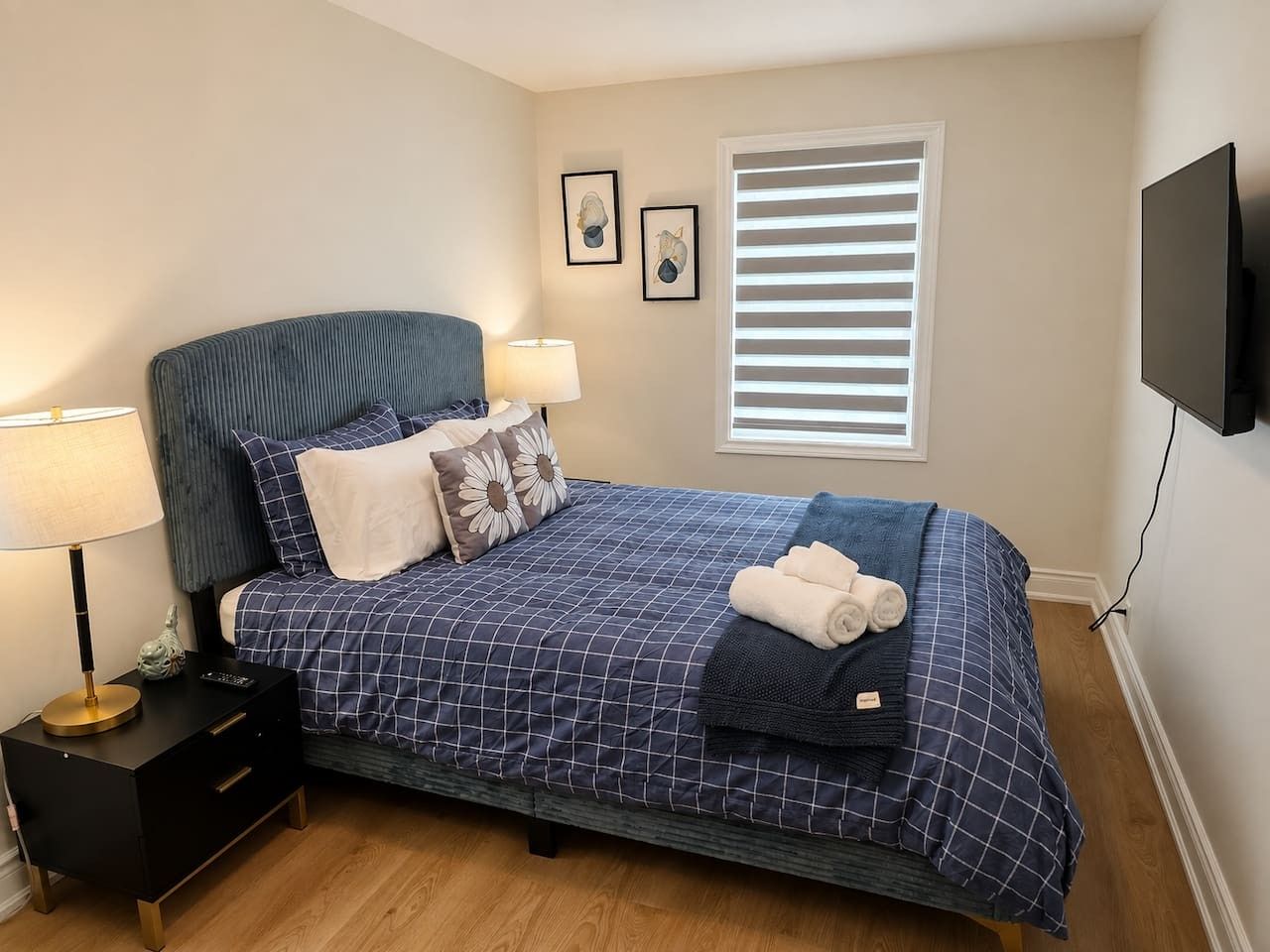 Une chambre à coucher comprenant un lit, des tables de chevet, une lampe, une fenêtre avec des stores et un téléviseur fixé au mur.