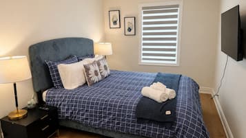 Une chambre à coucher comprenant un lit, des tables de chevet, une lampe, une fenêtre avec des stores et un téléviseur fixé au mur.