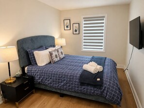 Une chambre à coucher comprenant un lit, des tables de chevet, une lampe, une fenêtre avec des stores et un téléviseur fixé au mur.