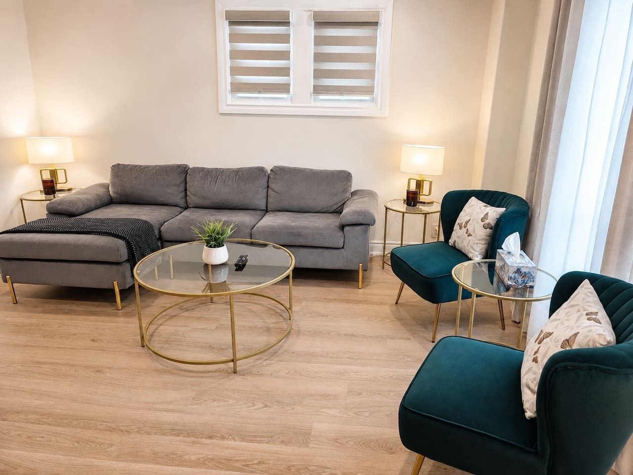 Un salon moderne avec un canapé gris, deux fauteuils, une table basse en verre et deux petites tables d’appoint avec des lampes.