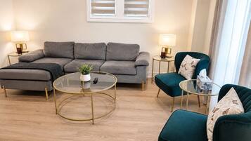 Un salon moderne avec un canapé gris, deux fauteuils, une table basse en verre et deux petites tables d’appoint avec des lampes.
