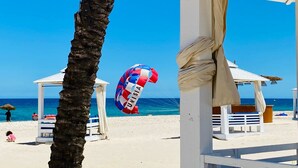 Escena de playa con cielo despejado, una palmera, sombrillas y una silla de playa.