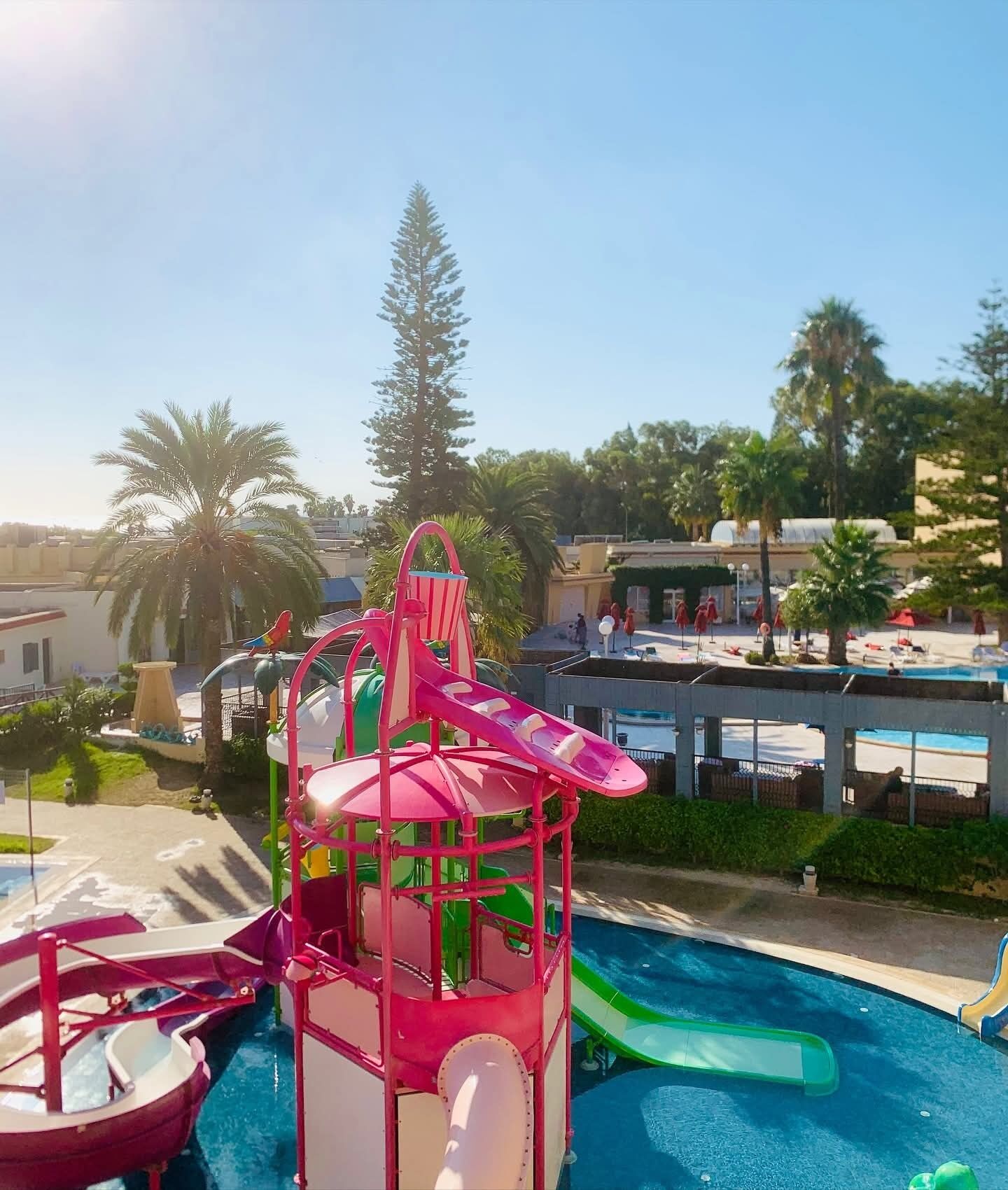Un parque acuático con una estructura central de juegos, piscina y palmeras a su alrededor.