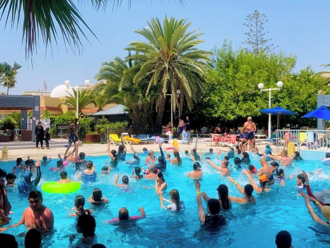 A swimming pool area with people swimming and playing, surrounded by palm trees and umbrellas.